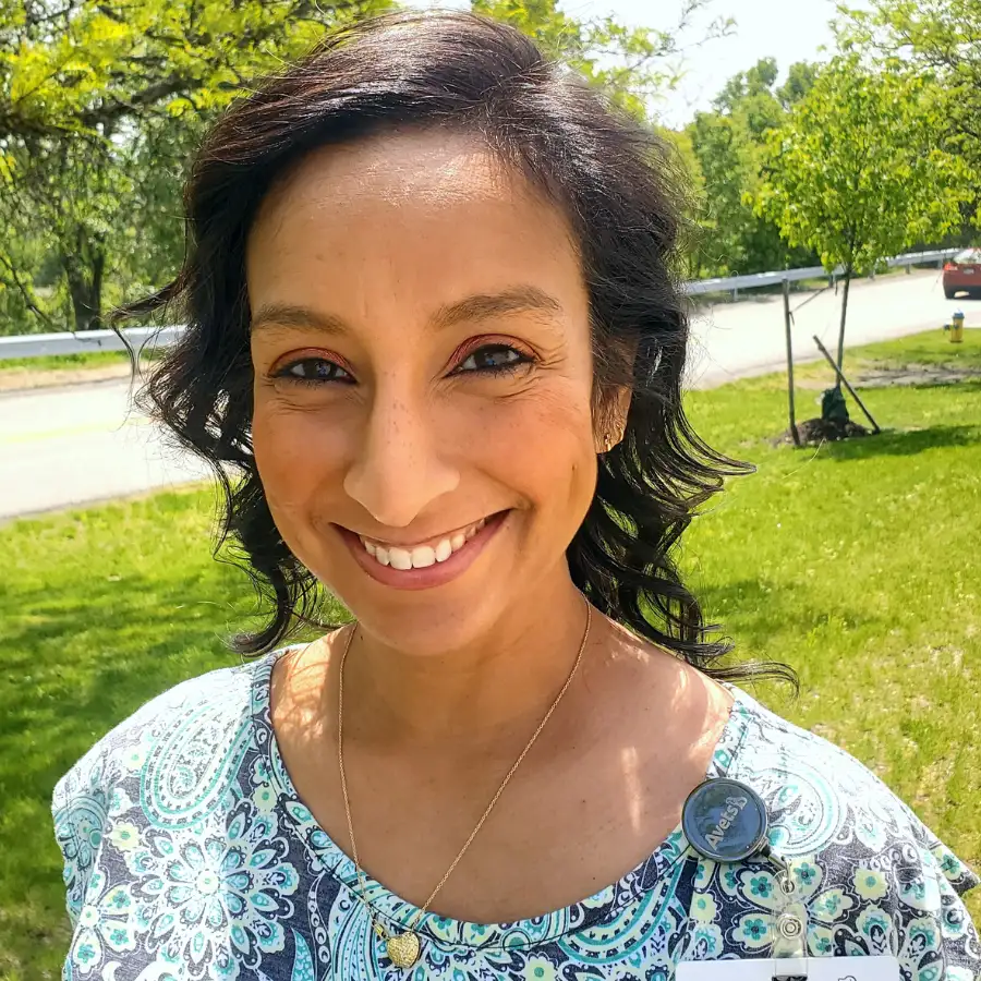 Smiling woman with medium-length wavy hair, wearing a patterned blue and white top, standing outdoors on a sunny day with a green grassy background.