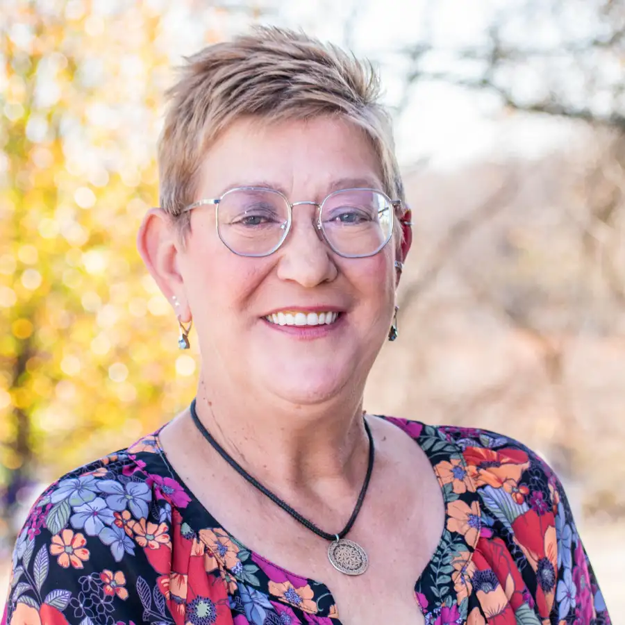 Smiling woman with short blonde hair, wearing glasses and a colorful floral top, standing outdoors on a sunny day with autumn foliage in the background.